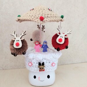 May include: A white ceramic mug with a smiling face, decorated with a white crocheted hat and a crocheted reindeer on top. The hat is adorned with candy cane buttons. Three reindeer ornaments hang from a crocheted beige umbrella on top of the mug.