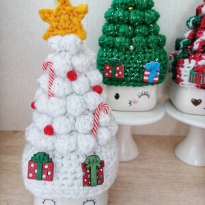 May include: Three white ceramic mugs with crocheted Christmas tree hats. The mugs have painted eyes and smiles. The trees are decorated with red and white candy canes, red beads, and small wooden presents.