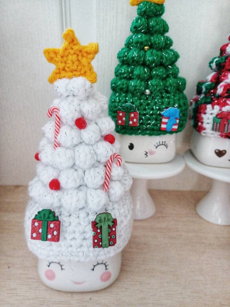 May include: Three white ceramic mugs with crocheted Christmas tree hats. The mugs have painted eyes and smiles. The trees are decorated with red and white candy canes, red beads, and small wooden presents.