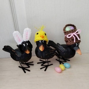 K&ouml;nnte beinhalten: Drei schwarze Vogelfiguren mit Osterh&uuml;ten. Ein Vogel tr&auml;gt Hasenohren, ein anderer einen K&uuml;kenhut und der dritte einen Korbhut. Pastellfarbene Ostereier sind in der N&auml;he der V&ouml;gel verstreut und schaffen eine festliche Fr&uuml;hlingsausstellung.