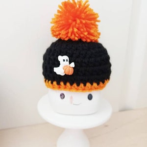 K&ouml;nnte beinhalten: Ein schwarzer geh&auml;kelter Hut mit einem orangefarbenen Bommel und einem wei&szlig;en Geisterknopf, der einen K&uuml;rbis h&auml;lt. Der Hut ist auf einem wei&szlig;en Keramik-Marshmallow mit einem l&auml;chelnden Gesicht platziert. Halloween-Dekoration.