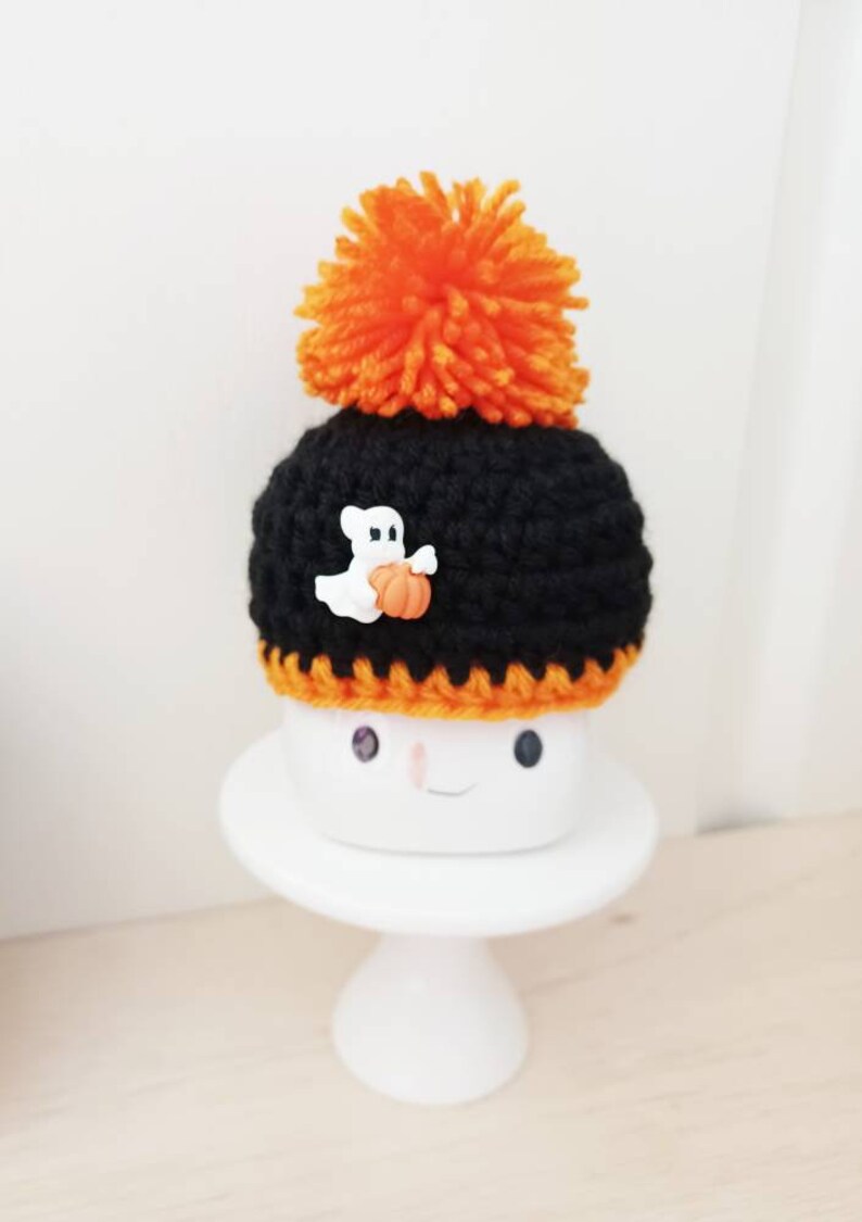 K&ouml;nnte beinhalten: Ein schwarzer geh&auml;kelter Hut mit einem orangefarbenen Bommel und einem wei&szlig;en Geisterknopf, der einen K&uuml;rbis h&auml;lt. Der Hut ist auf einem wei&szlig;en Keramik-Marshmallow mit einem l&auml;chelnden Gesicht platziert. Halloween-Dekoration.
