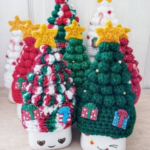 May include: Five crocheted Christmas trees in different colors and patterns, each with a yellow star on top. The trees are sitting on top of white mugs with cute faces.