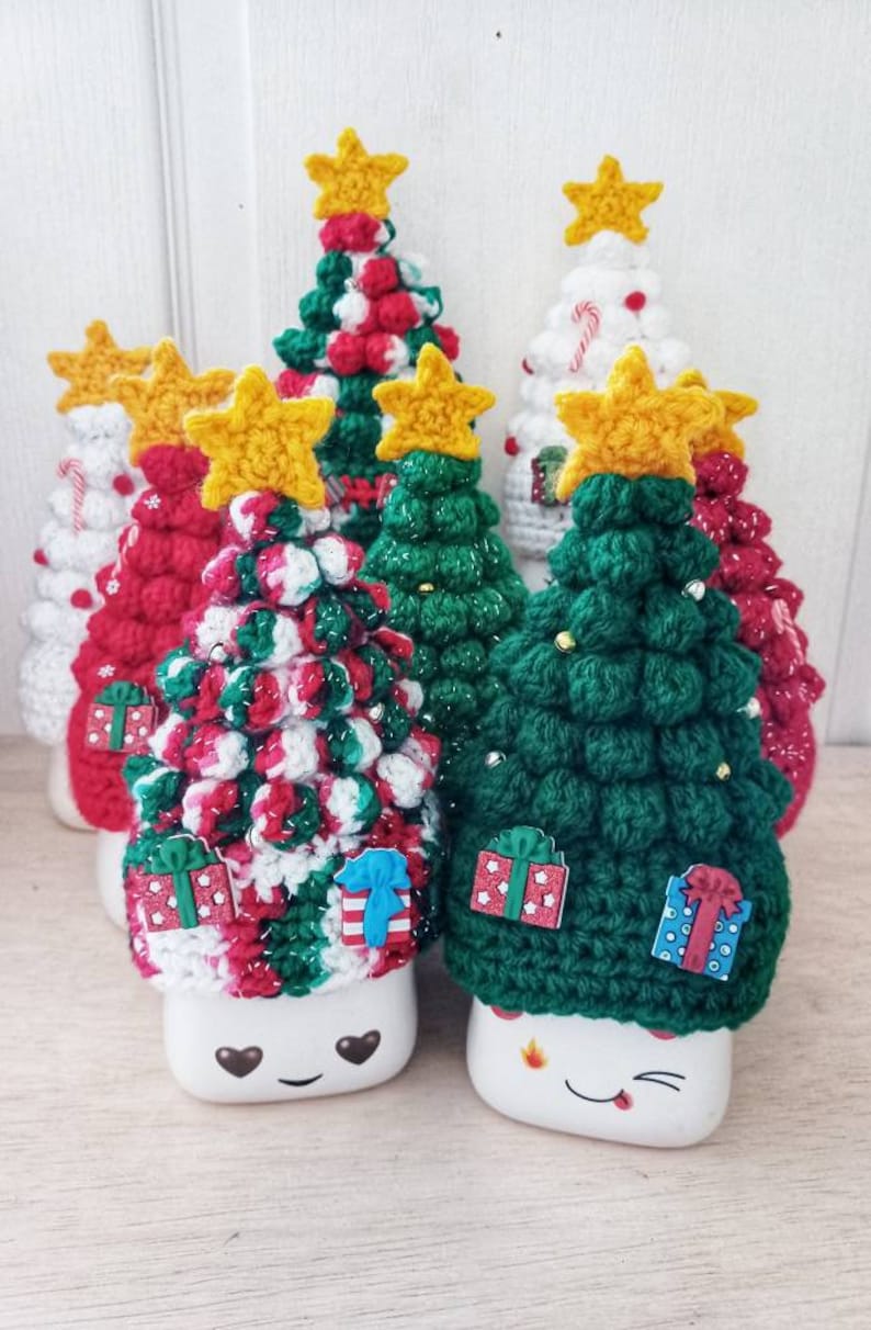 May include: Five crocheted Christmas trees in different colors and patterns, each with a yellow star on top. The trees are sitting on top of white mugs with cute faces.