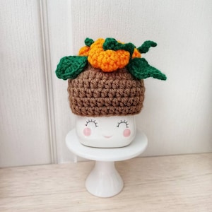 May include: A white ceramic mug with a smiling face, wearing a brown crocheted hat with orange and green crocheted pumpkins on top. The mug is sitting on a white cake stand.
