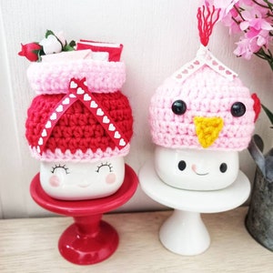 May include: Two ceramic mugs with crocheted hats. The mug on the left has a pink and red hat with white hearts. The mug on the right has a pink hat with a yellow beak and black eyes. Both mugs have a smiling face.