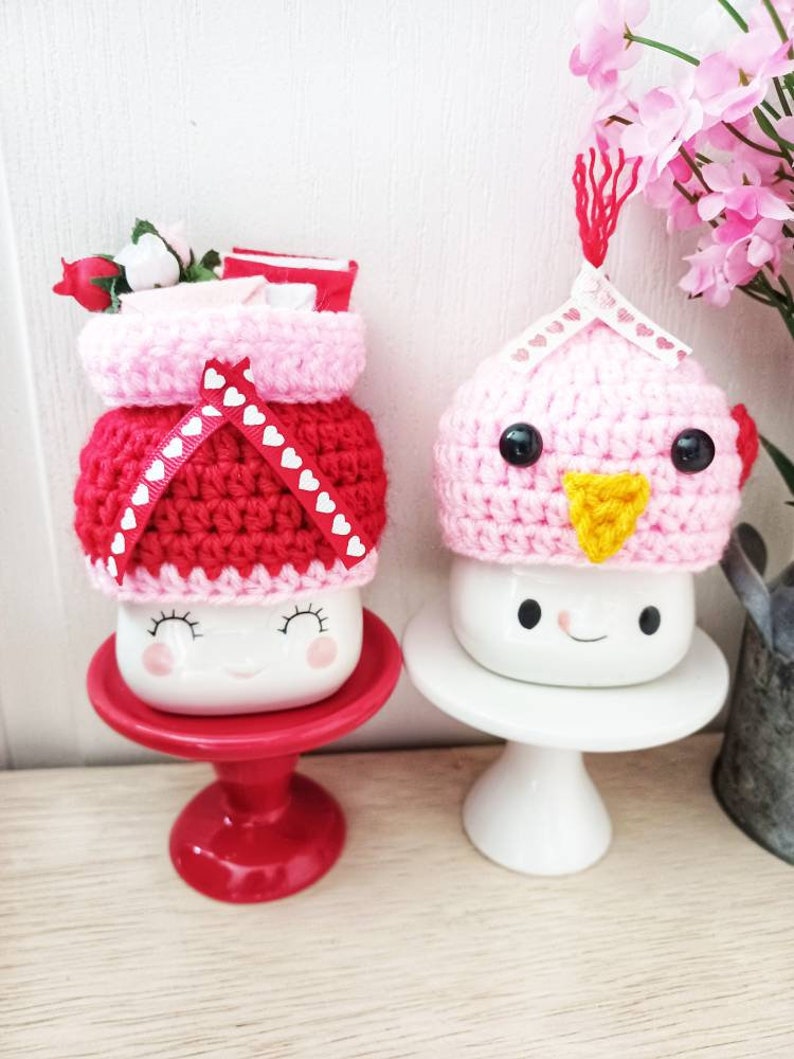 May include: Two ceramic mugs with crocheted hats. The mug on the left has a pink and red hat with white hearts. The mug on the right has a pink hat with a yellow beak and black eyes. Both mugs have a smiling face.