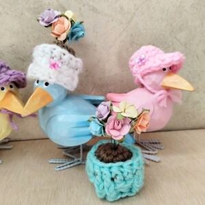 Op de afbeelding: Drie houten vogelbeeldjes met gehaakte hoeden en een kleine gehaakte bloempot met een bloemstuk. De vogels zijn geschilderd in lichtblauw, roze en geel. De hoeden zijn wit, roze en paars. De bloempot is turquoise.
