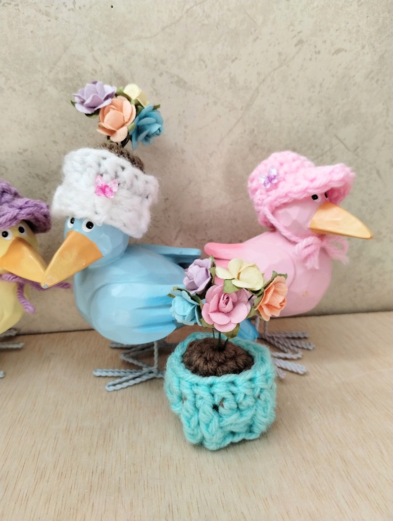 Op de afbeelding: Drie houten vogelbeeldjes met gehaakte hoeden en een kleine gehaakte bloempot met een bloemstuk. De vogels zijn geschilderd in lichtblauw, roze en geel. De hoeden zijn wit, roze en paars. De bloempot is turquoise.