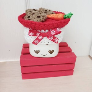 Op de afbeelding: Een witte keramische mok met een rood-witte gehaakte muts, een wortelneus en chocoladekoekjes. De mok heeft een glimlachend gezicht met hartvormige ogen. De mok staat op een rode houten kist.