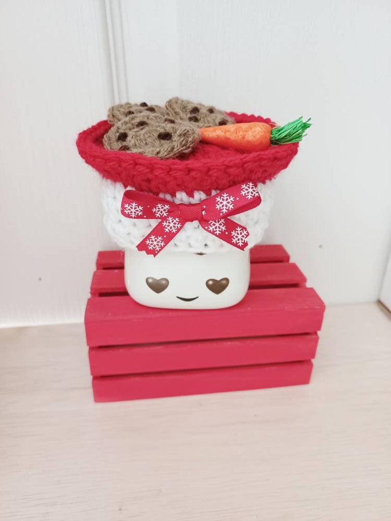 Op de afbeelding: Een witte keramische mok met een rood-witte gehaakte muts, een wortelneus en chocoladekoekjes. De mok heeft een glimlachend gezicht met hartvormige ogen. De mok staat op een rode houten kist.