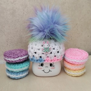 May include: A white crocheted hat with a rainbow colored pom pom and a small bunny charm. The hat is sitting on a white mug with a smiley face. There are three stacks of crocheted circles in pink, yellow, and blue.