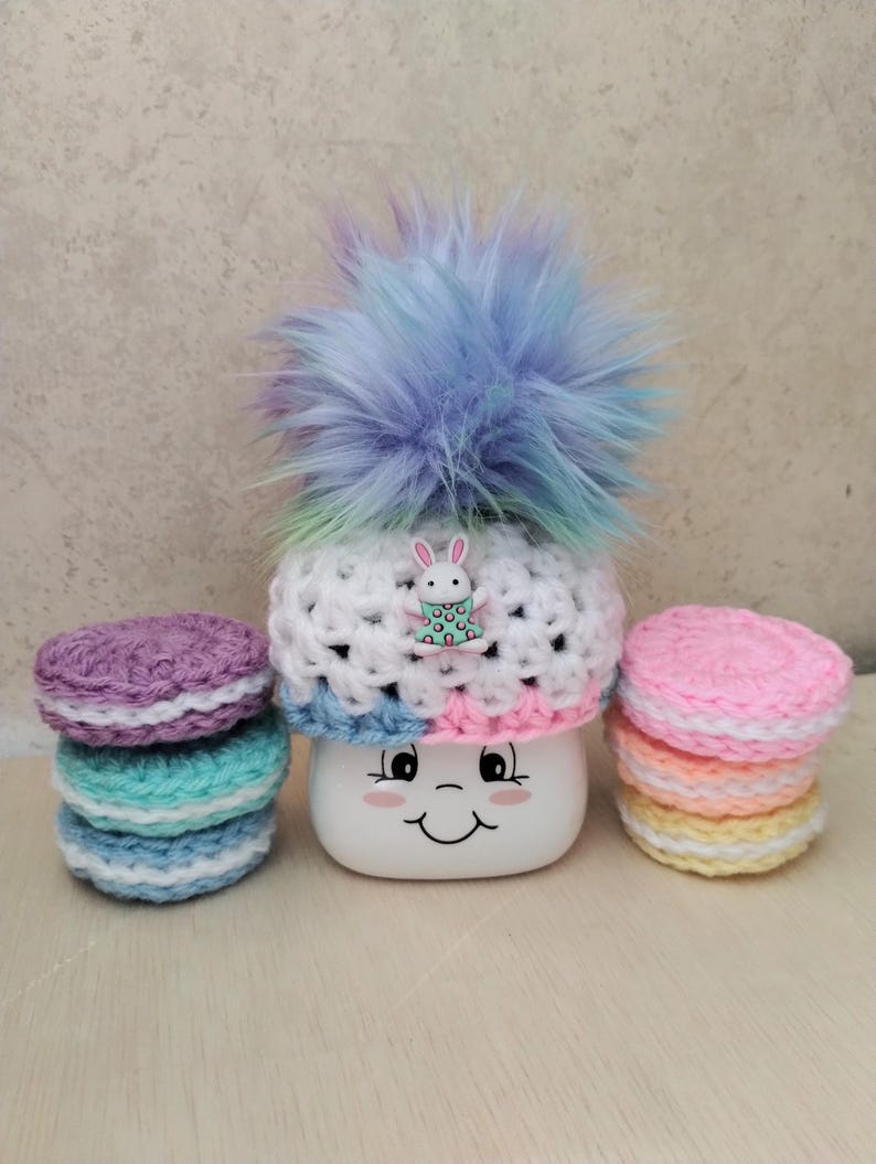 May include: A white crocheted hat with a rainbow colored pom pom and a small bunny charm. The hat is sitting on a white mug with a smiley face. There are three stacks of crocheted circles in pink, yellow, and blue.