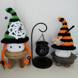 May include: Two crocheted witch hats with marshmallow faces and orange yarn hair. The hats are decorated with black and white stripes, a spider web, and a broom. The hats are sitting on small wooden stools. A black cauldron hangs from a metal stand.