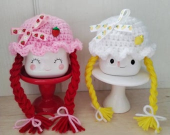 May include: Two white ceramic mugs with crocheted hats and yarn braids. One mug has a pink hat with strawberry print and red braids. The other mug has a white hat with lemon print and yellow braids.