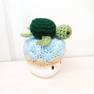 May include: A crocheted sea turtle with a green shell and light green flippers sits on top of a blue and tan crocheted hat. The hat is on a white mug with a smiling face.
