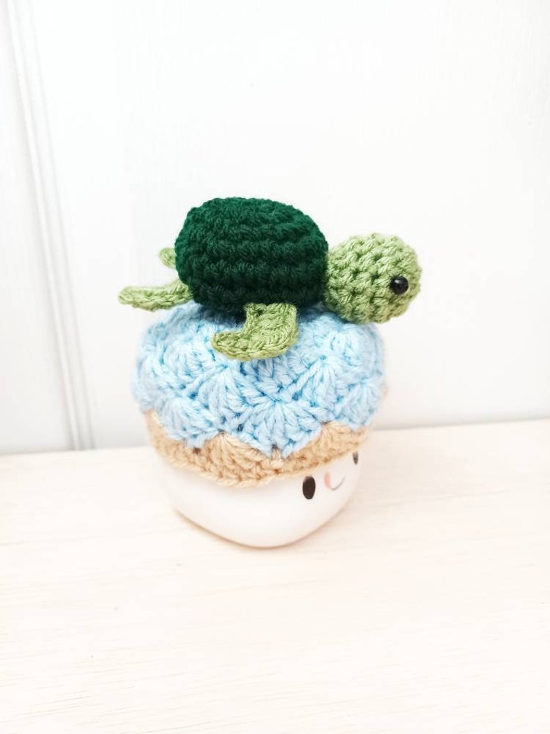 May include: A crocheted sea turtle with a green shell and light green flippers sits on top of a blue and tan crocheted hat. The hat is on a white mug with a smiling face.