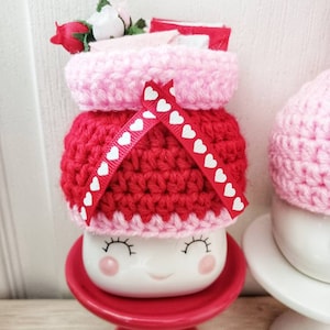 May include: A white ceramic mug with a smiling face, wearing a red and pink crocheted hat with white heart accents. The mug is sitting on a red pedestal.