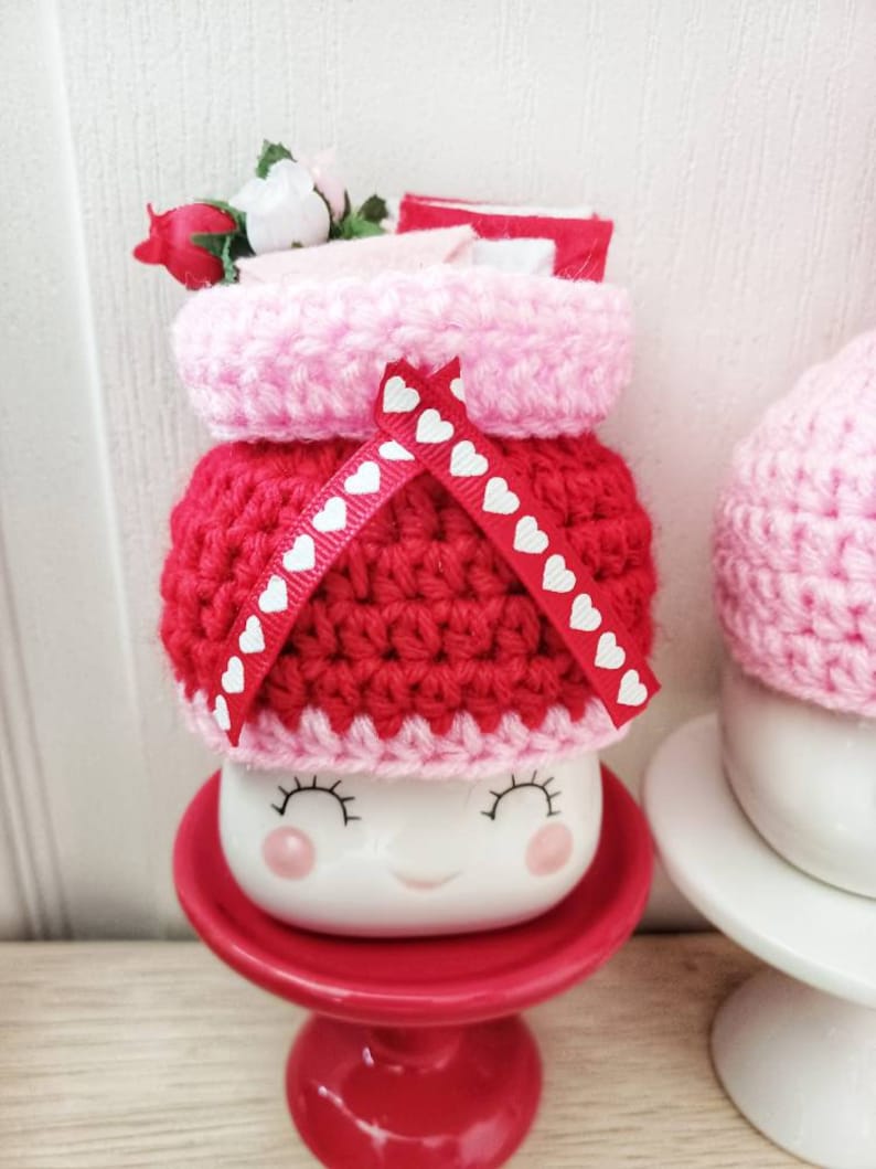 May include: A white ceramic mug with a smiling face, wearing a red and pink crocheted hat with white heart accents. The mug is sitting on a red pedestal.