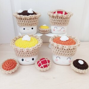 May include: Four white ceramic mugs with crocheted pie toppers. Each mug has a happy face and is topped with a different pie: chocolate, pumpkin, apple, and pecan. There are also four smaller crocheted pies on the table.