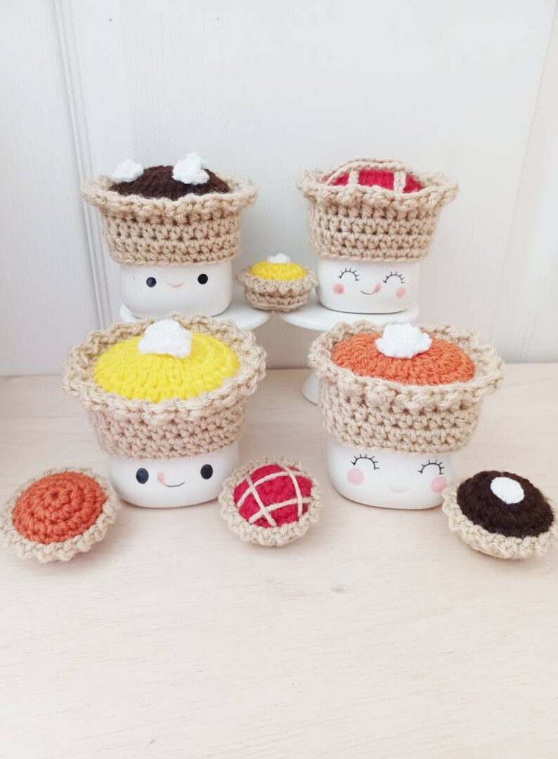 May include: Four white ceramic mugs with crocheted pie toppers. Each mug has a happy face and is topped with a different pie: chocolate, pumpkin, apple, and pecan. There are also four smaller crocheted pies on the table.