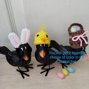 K&ouml;nnte beinhalten: Drei schwarze Vogelfiguren mit Osterh&uuml;ten. Ein Vogel tr&auml;gt Hasenohren, ein anderer einen K&uuml;kenhut und der dritte einen Korbhut. Pastellfarbene Eier sind darunter verstreut. Die V&ouml;gel haben orangefarbene Schn&auml;bel und Drahtbeine.
