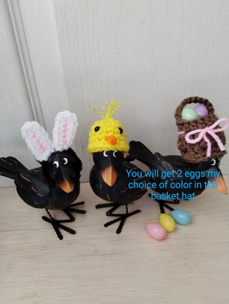 K&ouml;nnte beinhalten: Drei schwarze Vogelfiguren mit Osterh&uuml;ten. Ein Vogel tr&auml;gt Hasenohren, ein anderer einen K&uuml;kenhut und der dritte einen Korbhut. Pastellfarbene Eier sind darunter verstreut. Die V&ouml;gel haben orangefarbene Schn&auml;bel und Drahtbeine.