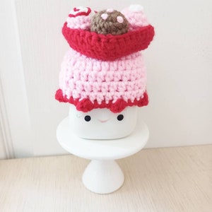May include: A pink and red crocheted cake with a brown chocolate topping and white frosting. The cake is sitting on a white cake stand.