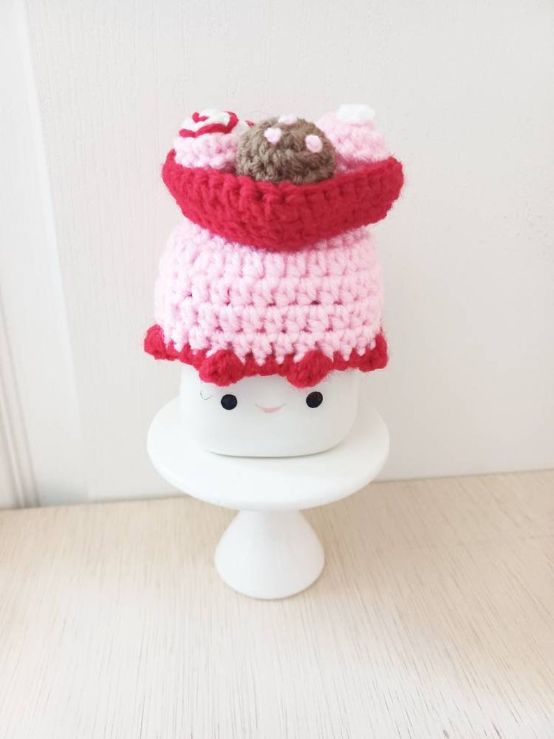 May include: A pink and red crocheted cake with a brown chocolate topping and white frosting. The cake is sitting on a white cake stand.