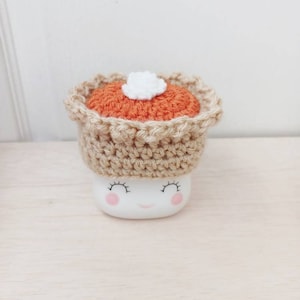May include: A white ceramic mug with a smiling face and pink cheeks, wearing a crocheted pumpkin pie hat with a white dollop of whipped cream on top.