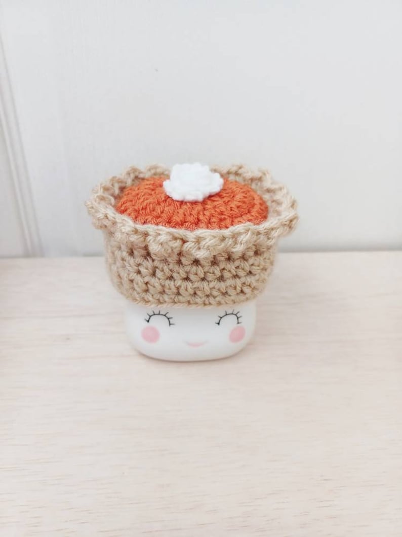 May include: A white ceramic mug with a smiling face and pink cheeks, wearing a crocheted pumpkin pie hat with a white dollop of whipped cream on top.