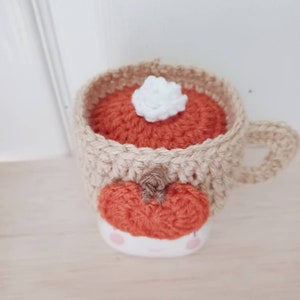 May include: A crocheted mug cozy in the shape of a pumpkin spice latte. The cozy is beige with an orange pumpkin and a white marshmallow on top.