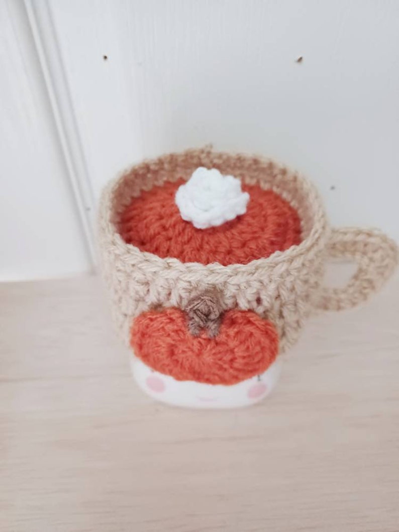 May include: A crocheted mug cozy in the shape of a pumpkin spice latte. The cozy is beige with an orange pumpkin and a white marshmallow on top.