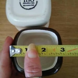 May include: Two square ceramic ramekins, one white with black lettering and one brown with a white interior. The white ramekin has the text "One Hundred 80 degrees Dishwasher Safe Microwave Safe" and the number "180" in a circle. A yellow measuring tape is wrapped around the brown ramekin, measuring 3 inches.