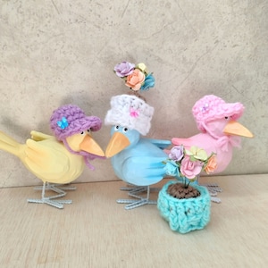 May include: Three wooden bird figurines with crocheted hats and flowers. The birds are yellow, blue, and pink. The blue bird is holding a small crocheted flower pot with flowers.