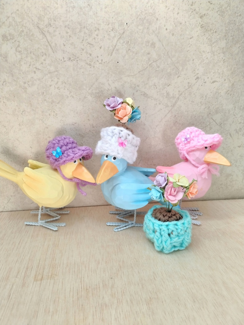 Op de afbeelding: Drie houten vogelbeeldjes met gehaakte hoeden en bloemen. De vogels zijn geel, blauw en roze. De blauwe vogel houdt een klein gehaakt bloempotje met bloemen vast.