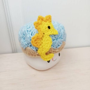 May include: A blue crocheted hat with a yellow crocheted seahorse on top. The hat is sitting on a white mug with a smiley face.