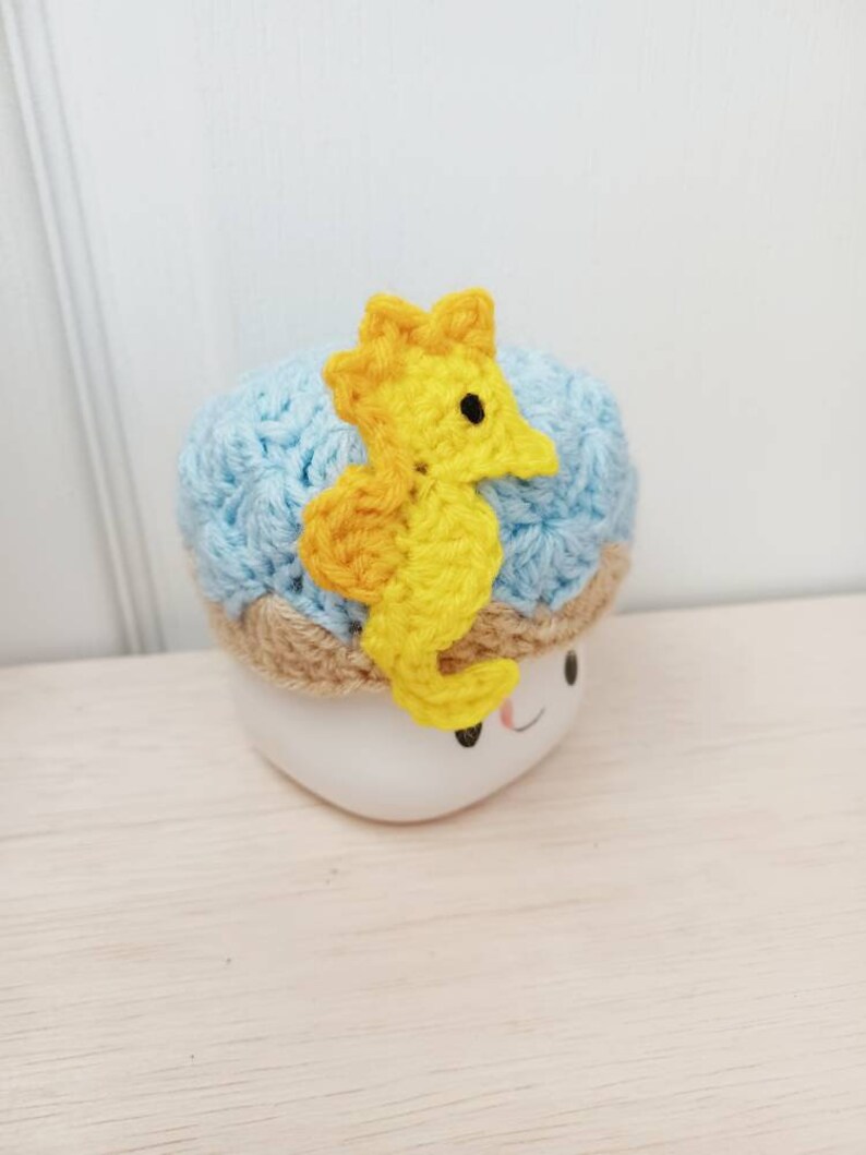 May include: A blue crocheted hat with a yellow crocheted seahorse on top. The hat is sitting on a white mug with a smiley face.