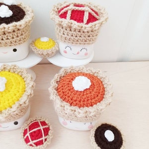 May include: Crocheted pie mug toppers in various colors and designs, including red, orange, yellow, and brown. The toppers are placed on top of white mugs with smiling faces. There are also smaller crocheted pie decorations on a wooden surface.