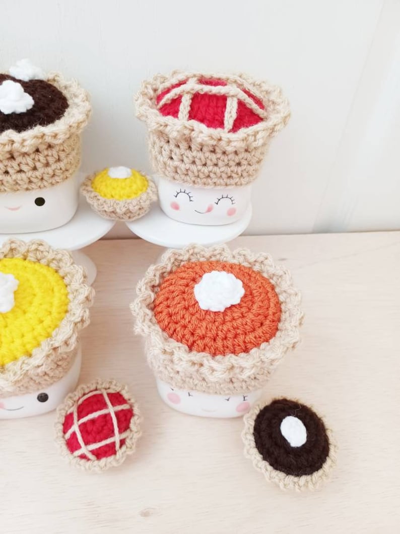 May include: Crocheted pie mug toppers in various colors and designs, including red, orange, yellow, and brown. The toppers are placed on top of white mugs with smiling faces. There are also smaller crocheted pie decorations on a wooden surface.