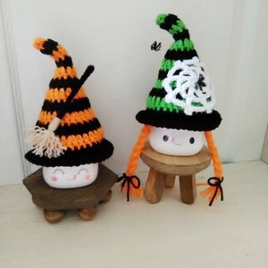 May include: Two white ceramic marshmallow figures dressed as witches for Halloween. One witch wears a black and orange striped hat with a broom and the other wears a green and black striped hat with a white crocheted spiderweb. Both witches have orange yarn hair.