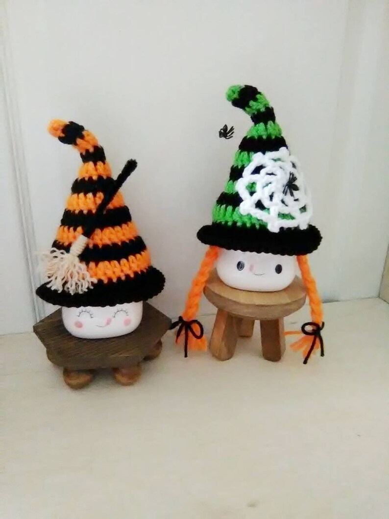 May include: Two white ceramic marshmallow figures dressed as witches for Halloween. One witch wears a black and orange striped hat with a broom and the other wears a green and black striped hat with a white crocheted spiderweb. Both witches have orange yarn hair.