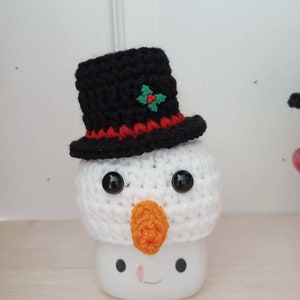 May include: A white crocheted snowman with a black top hat, red trim, and a green holly accent. The snowman has black eyes, an orange nose, and a white mug with a smiling face.