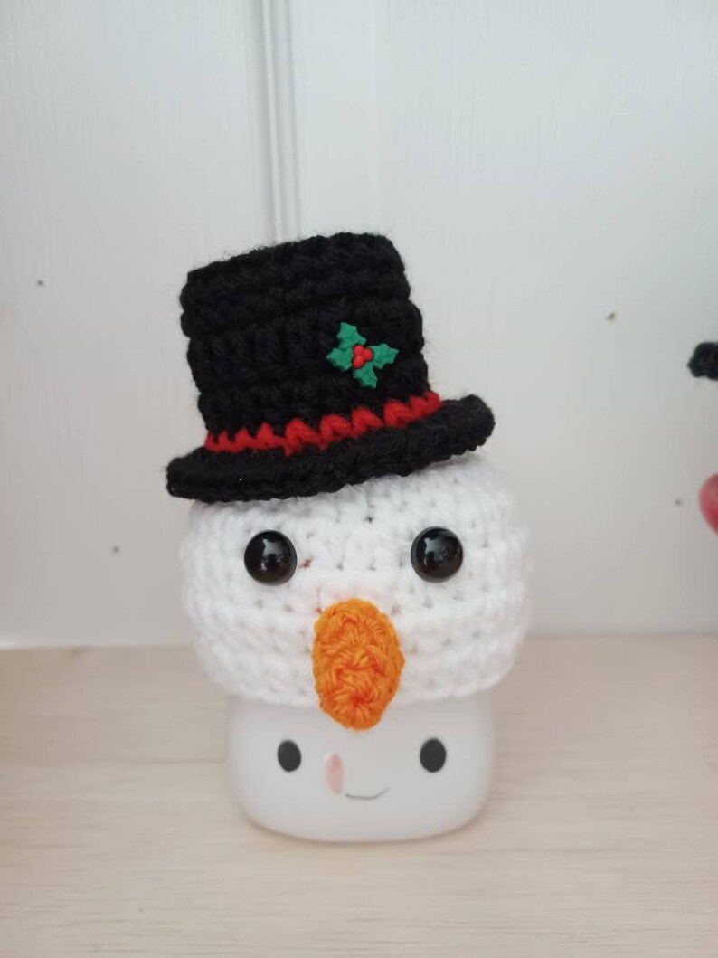 May include: A white crocheted snowman with a black top hat, red trim, and a green holly accent. The snowman has black eyes, an orange nose, and a white mug with a smiling face.