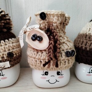 May include: Three white ceramic mugs with brown and beige crocheted hats. The mugs have a smiling face with pink cheeks and black eyes. The hats have brown coffee beans on them. The middle mug has a wooden spoon with coffee beans on it.