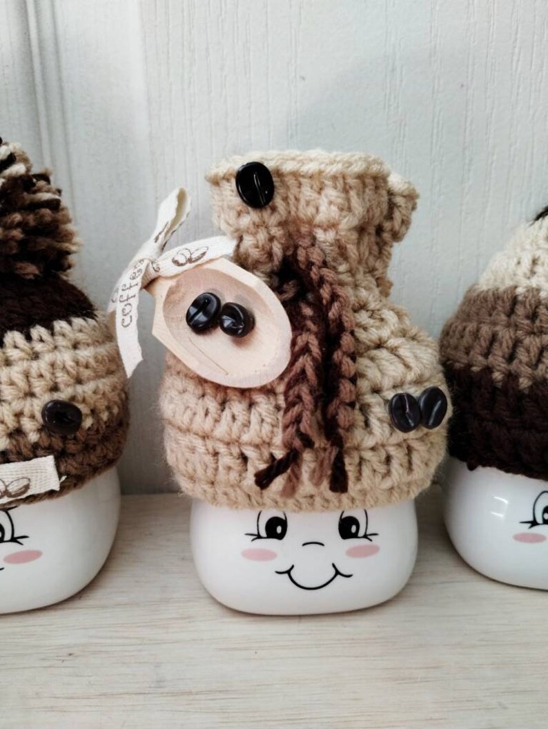 May include: Three white ceramic mugs with brown and beige crocheted hats. The mugs have a smiling face with pink cheeks and black eyes. The hats have brown coffee beans on them. The middle mug has a wooden spoon with coffee beans on it.