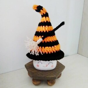 May include: A white ceramic mug with a smiling face, wearing a black and orange striped crocheted witch hat with a white broom.