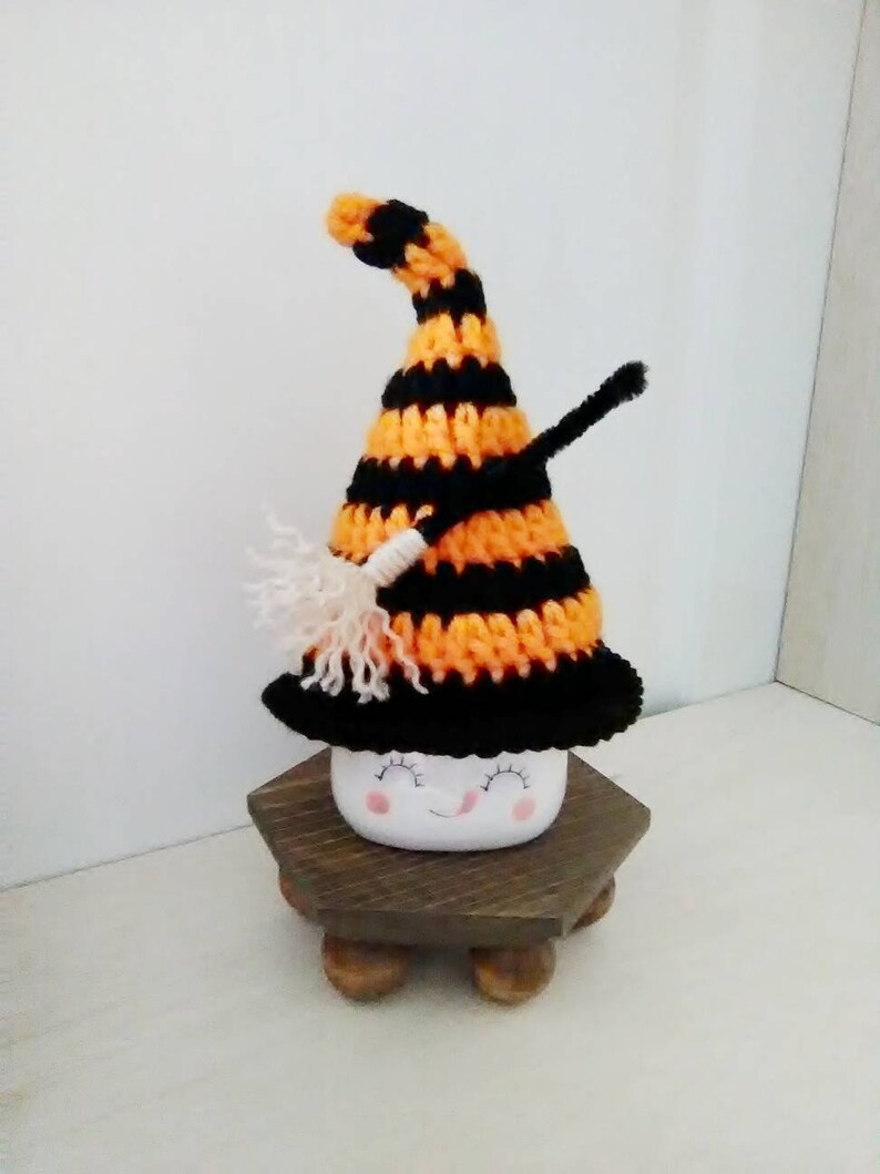 May include: A white ceramic mug with a smiling face, wearing a black and orange striped crocheted witch hat with a white broom.