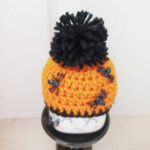 K&ouml;nnte beinhalten: Eine orangefarbene H&auml;kelm&uuml;tze mit einem gro&szlig;en schwarzen Bommel und drei kleinen schwarzen Spinnen. Die M&uuml;tze sitzt auf einem wei&szlig;en Keramikgesicht mit geschlossenen Augen und einem l&auml;chelnden Mund, platziert auf einem schwarzen Sockel.