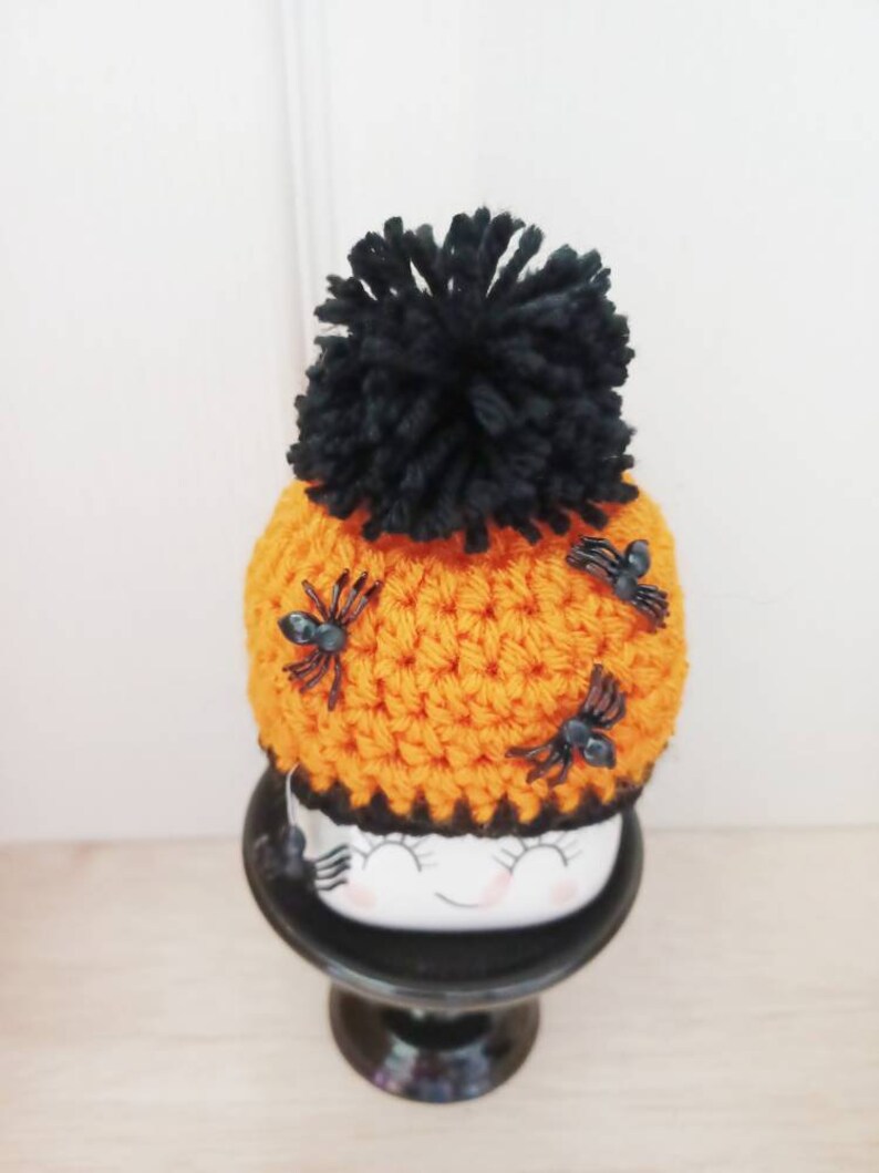K&ouml;nnte beinhalten: Eine orangefarbene H&auml;kelm&uuml;tze mit einem gro&szlig;en schwarzen Bommel und drei kleinen schwarzen Spinnen. Die M&uuml;tze sitzt auf einem wei&szlig;en Keramikgesicht mit geschlossenen Augen und einem l&auml;chelnden Mund, platziert auf einem schwarzen Sockel.